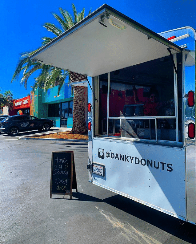 Modern food truck with a chalkboard menu, set against a sunny backdrop with palm trees and colorful shops.